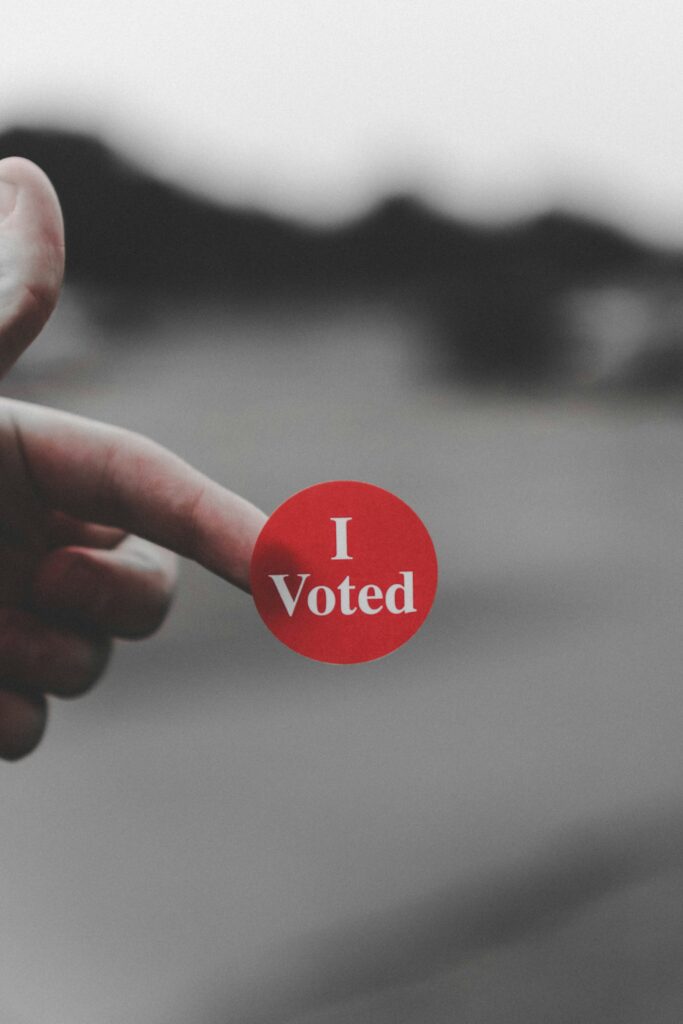 Hand holding a round red sticker reading 'I Voted' against a blurred background.