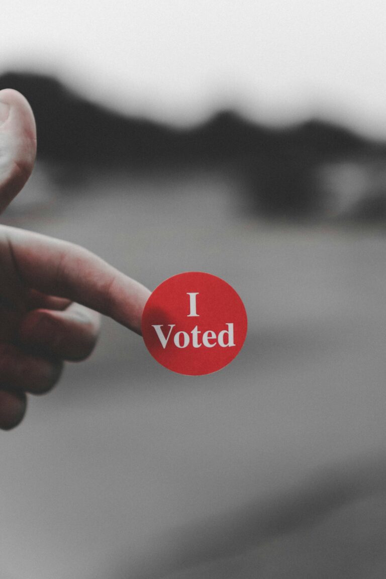 Hand holding a round red sticker reading 'I Voted' against a blurred background.